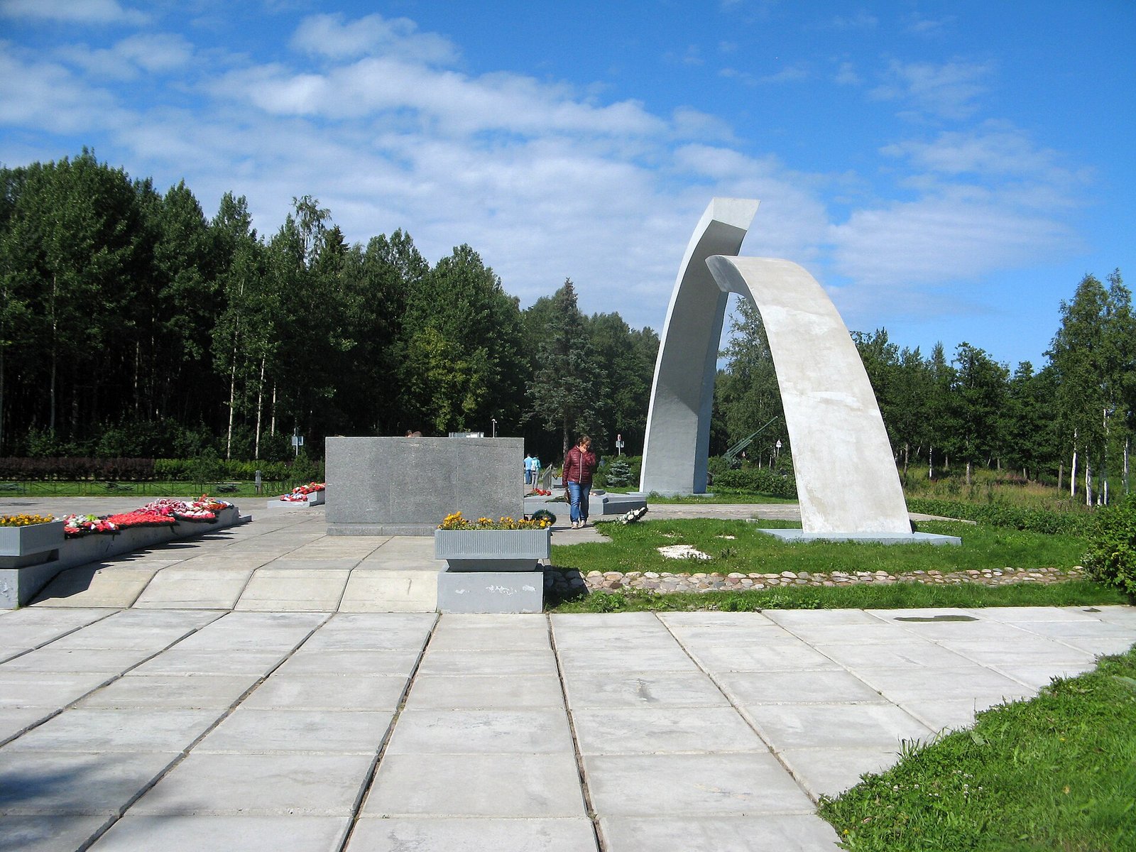 The Broken Ring memorial from the side: an arc, the square and flowers by the plinth